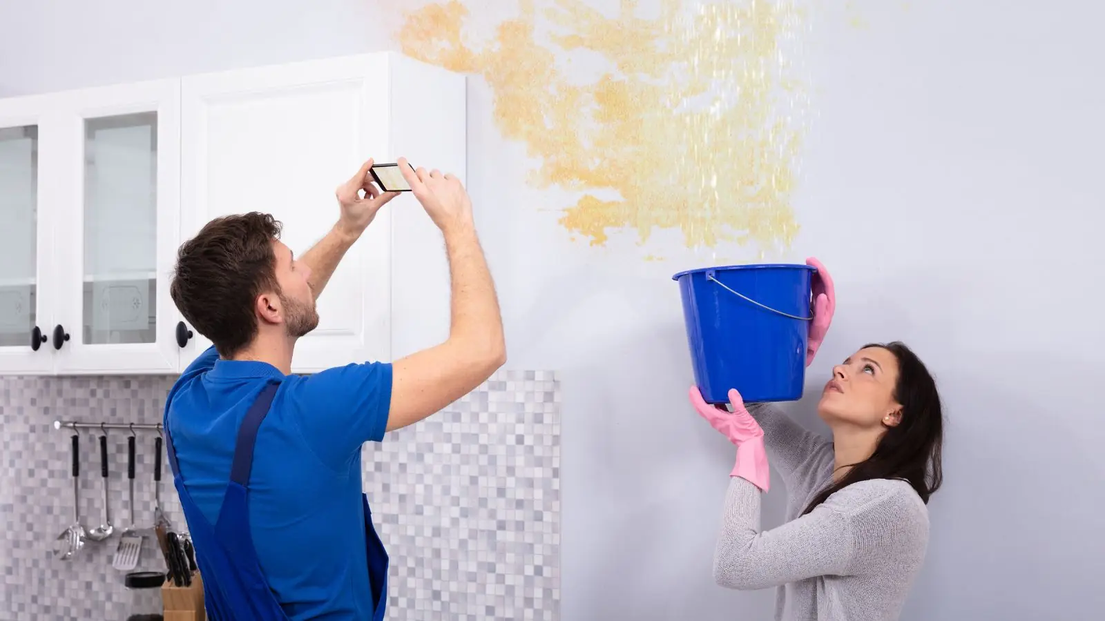Homeowner photographing a water-stained wall while another person holds a bucket under the leak